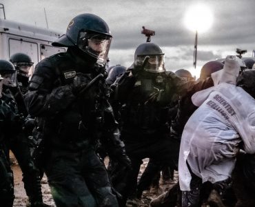 Ein Aktivist von Scientist Rebellion duckt sich weg um dem angedrohten Schlag einer Polizistin auszuweichen, die ihm gegenübersteht. Im Hintergrund sind der dunkle Himmel über Lützerath, einige Polizist:innen und ein Gefangenentransporter zu sehen.